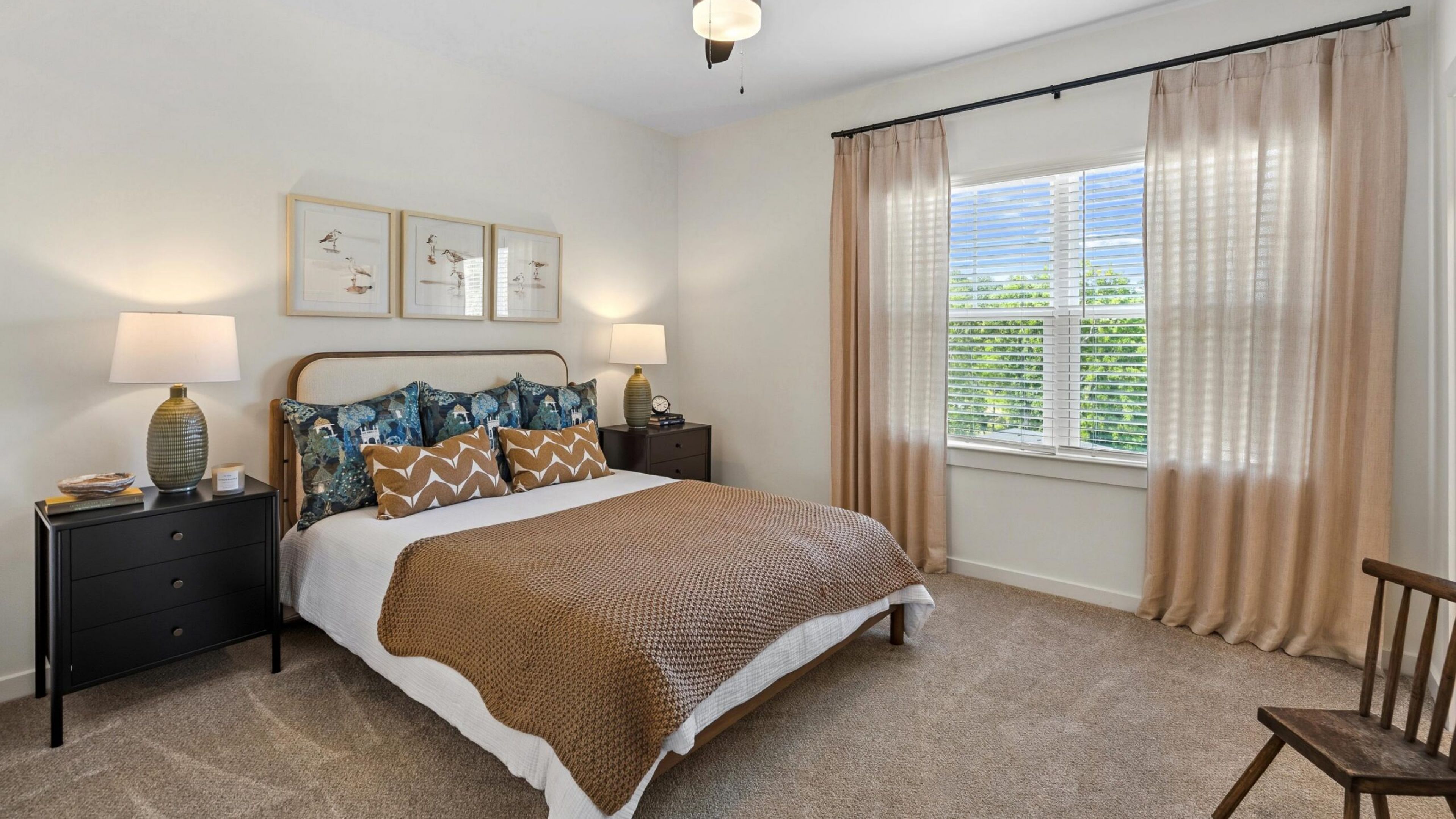 Cozy bedroom with a bed, patterned pillows, ceiling fan, framed art, and a window with beige curtains.