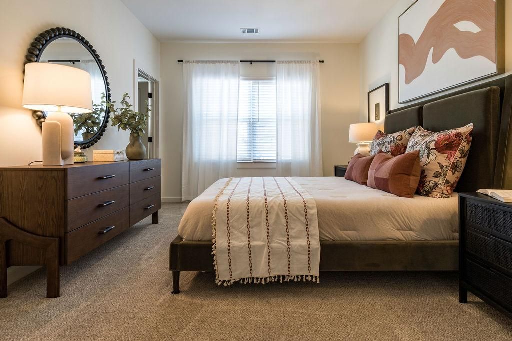 Cozy bedroom with a large bed, brown dresser, round mirror, and sunlight streaming through sheer curtains.