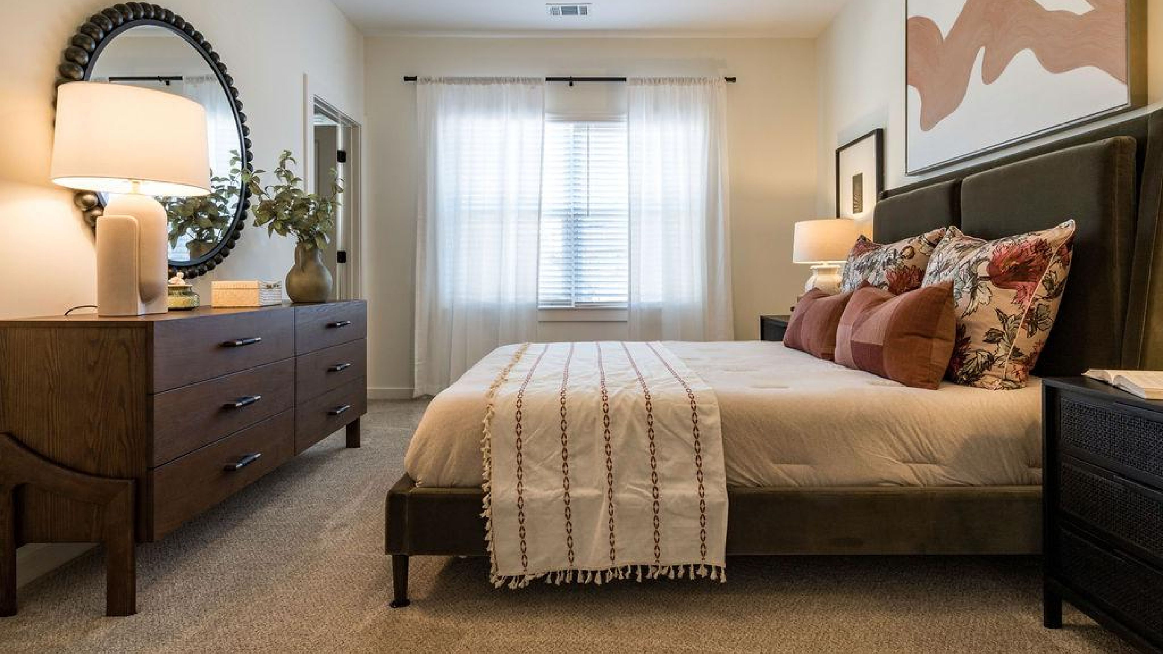 Cozy bedroom with a large bed, brown dresser, round mirror, and sunlight streaming through sheer curtains.